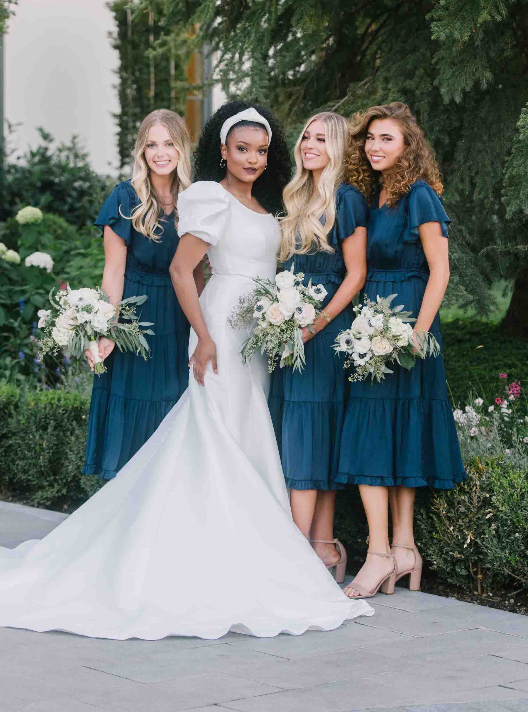 Bride wearing a mikado modest wedding dress with large pouf sleeves. She has three bridesmaids gathered around her all wearing the same navy modest bridesmaid dress from moment made shop.