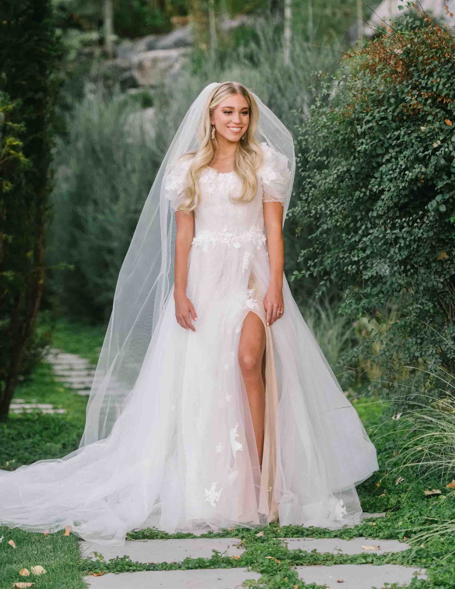 Bride standing along a stone pathway wearing a modest wedding dress with a slit above the know. This dress has a ballgown Silhouette with floral appliques and 