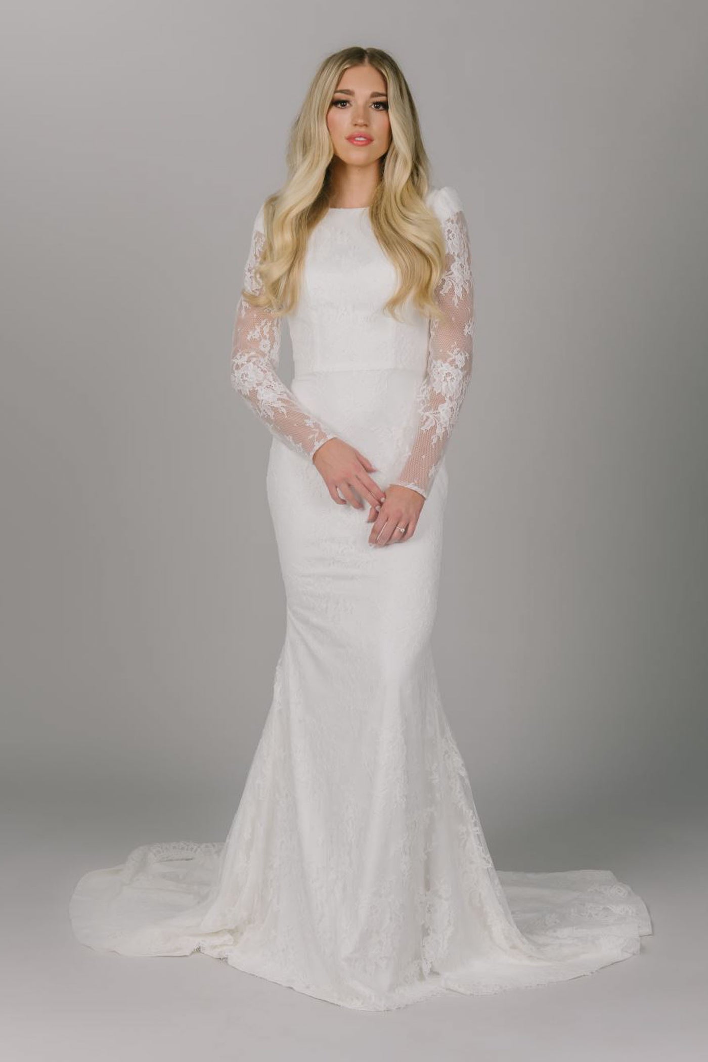 This is a blonde model modeling a modest wedding dress with full lace. It is long sleeved with a boat neckline and fitted silhouette.
