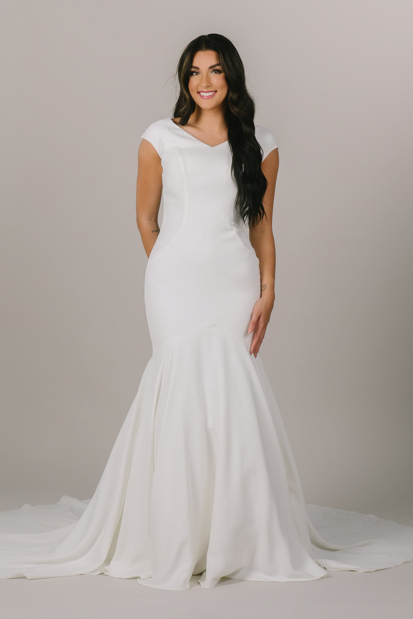 A brunette modeling standing with her arms to her side wearing a fit and flare modest wedding dress with a v neckline. 