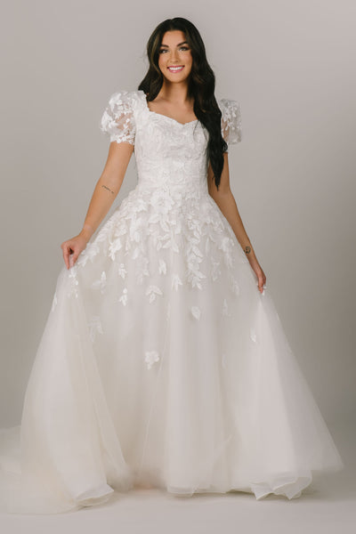 This is a front shot of a modest wedding dress with floral applique on the bodice and skirt, a-line silhouette, and puff sleeves.