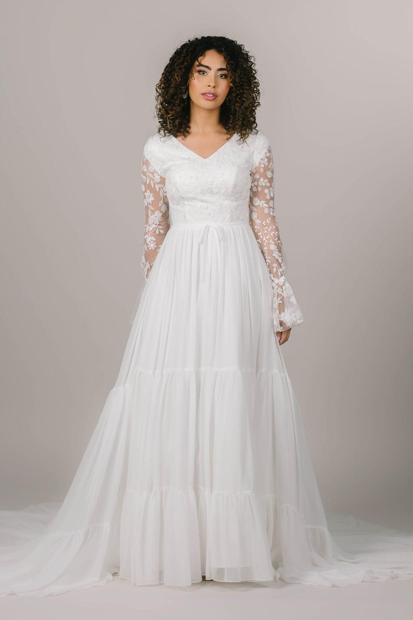 This is a modest wedding dress with a v neckline, sheer flowery sleeves, and a tiered a line skirt.