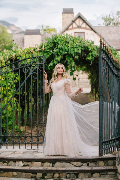A bride standing at a gate wearing a modest wedding dress blowing in the wind