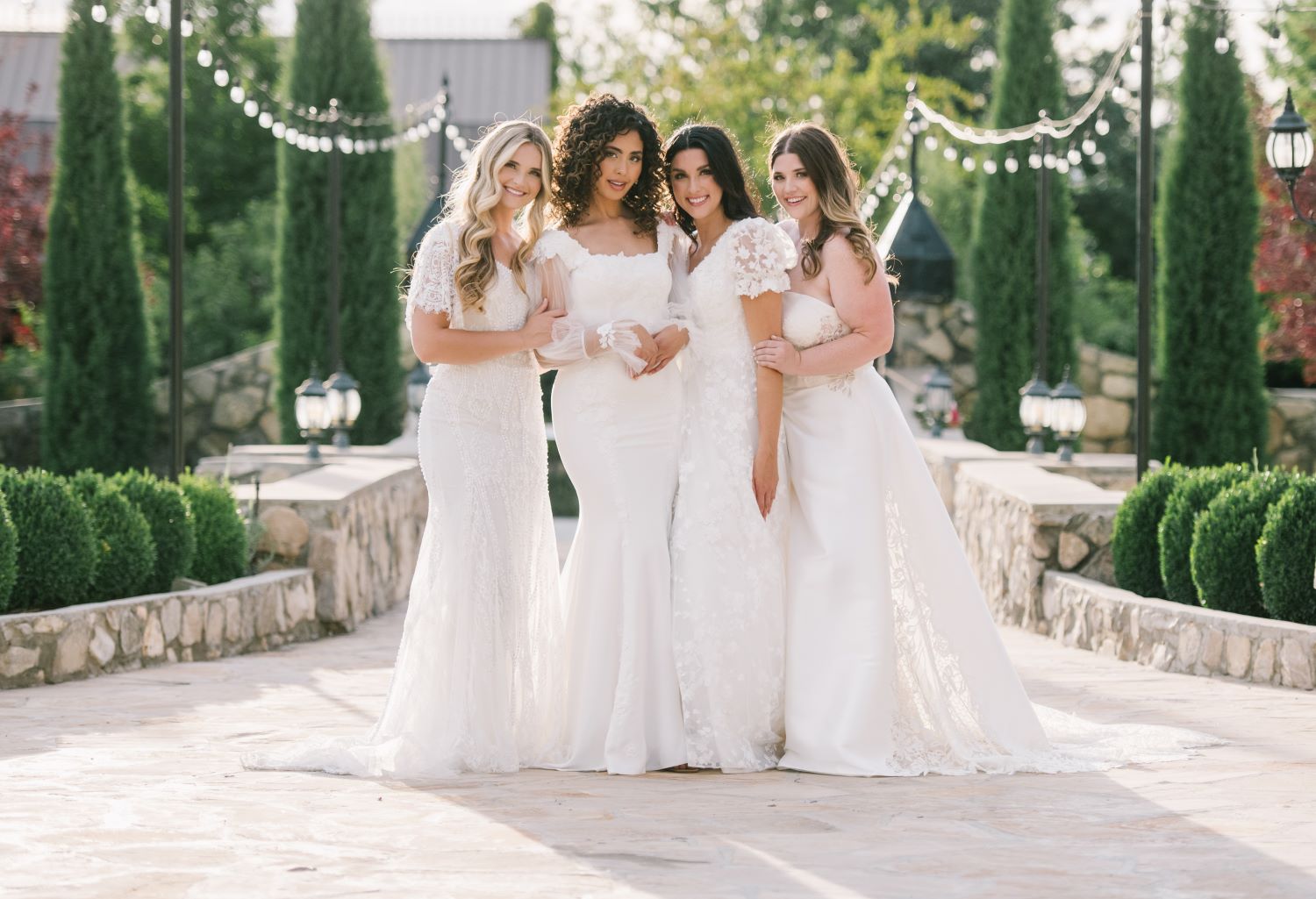 Four women wearing modest wedding dresses and strapless wedding dresses standing together on a stone path.