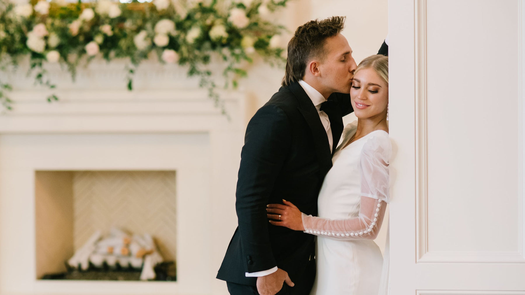 Groom kissing the forehead of a bride wearing a sheer long sleeve modest wedding dress. The sleeve is a pouf sleeve with button details down the side of the sleeve.