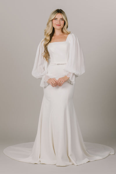 This is a modest wedding dress with a square neckline, billowy bishop sleeves, and a bow on the waistline.