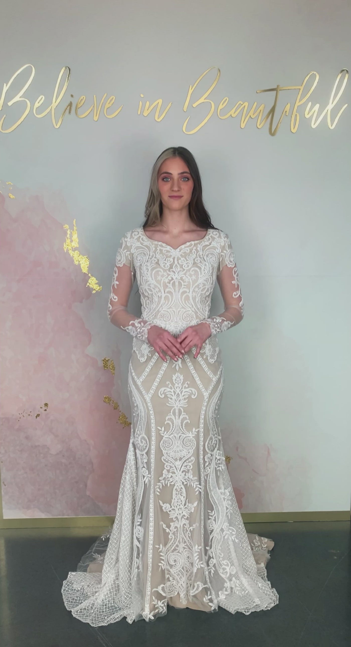 Video of modest wedding dress with long sleeves. It has a heart shaped neckline and fitted silhouette. It has lace detailing covering the whole dress. It has buttons on the sleeves.