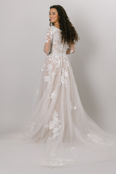 This modest wedding gown features long sleeves with lace decals throughout the bodice, skirt, and sleeves. It also has buttons running all the way down the train. 