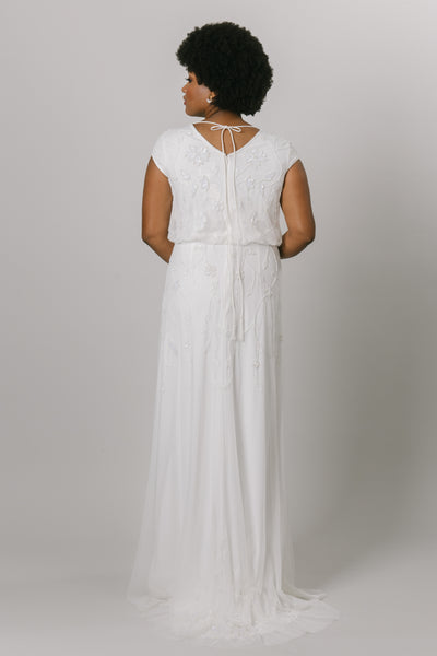 Back view of fitted modest wedding dress with cap sleeves. This dress has sheet lace and a fitted fit. It has a v-neckline with a lace scoop neckline overtop. It has a high tie at the neck. 