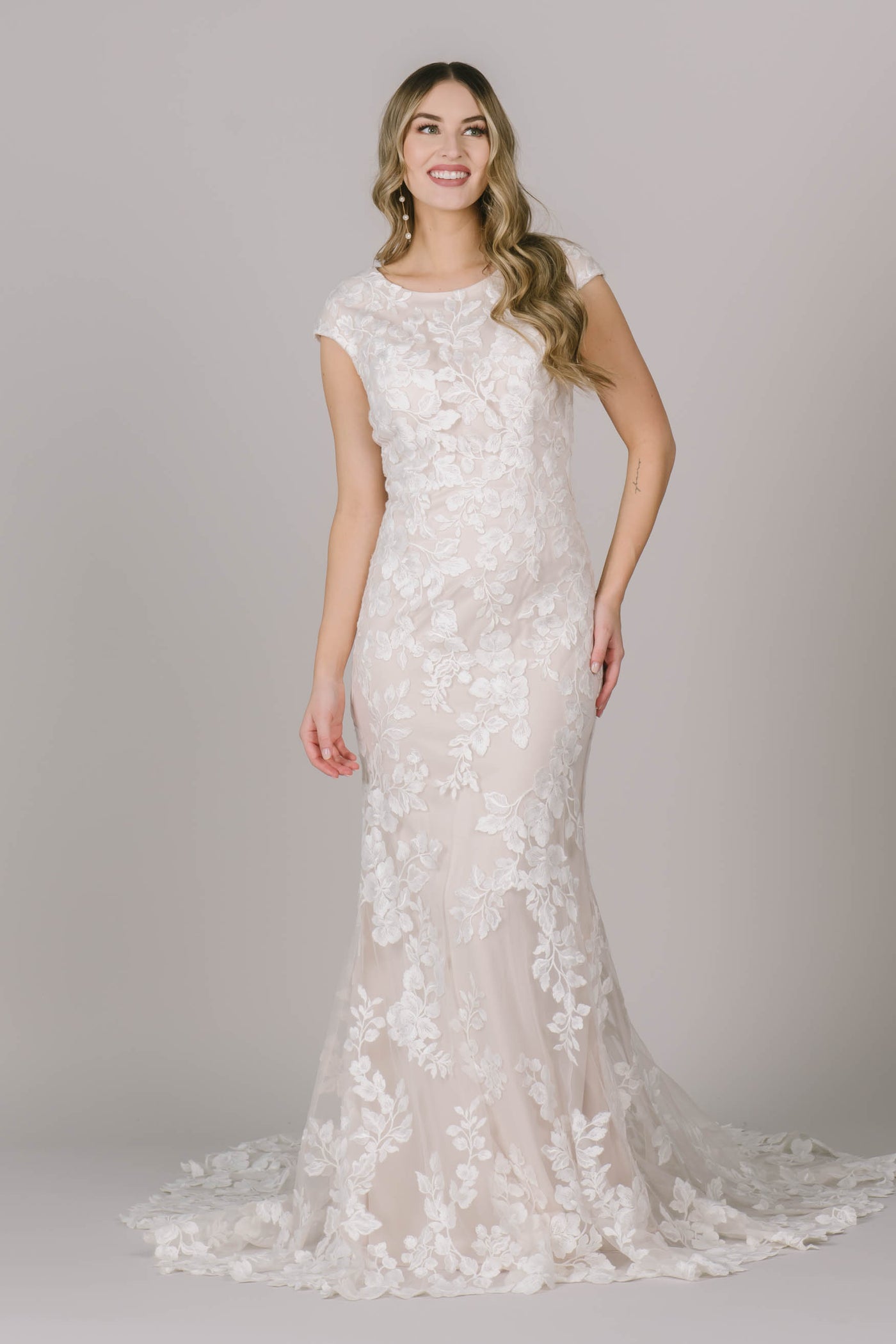 This fitted lace modest wedding dress features beautiful floral appliques, cap sleeves, and a higher scoop neckline.