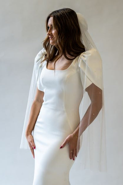 A front view portrait shot of a fingertip length bridal bow veil with pearls spread throughout hanging over the sleeves a wedding gown. 