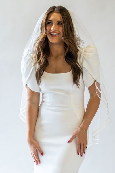 A front portrait shot of a fingertip length bridal veil with a double satin ribbon edge falling around a bride in a wedding gown.