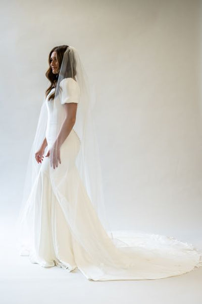 A side profile full body shot of a cathedral length veil with lace lining the bottom edge. The veil is spread out dramatically over the train of a simple modern wedding gown.