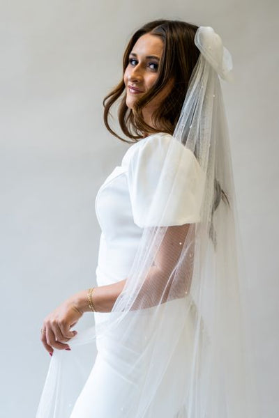 A side profile portrait shot of a cathedral length bow veil made of soft English netting containing pearls scattered throughout. 
