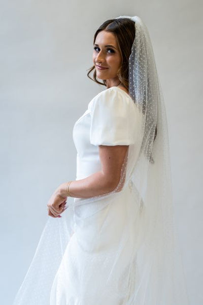 A side profile, portrait shot of a floor length, swiss dot bridal veil draping beautifully over the back and side of a wedding gown.