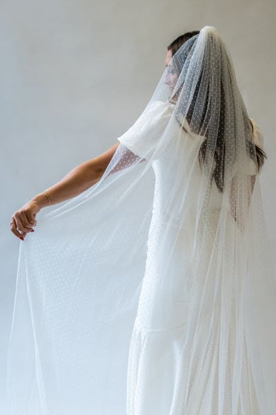 A back view portrait shot of a swiss dot floor length bridal veil being held out to the side to show the gorgeous pattern, perfect for adding a fun touch to any wedding gown.