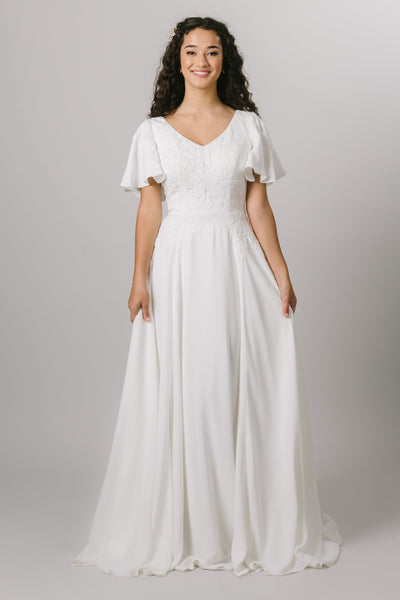 A simple and flowy modest wedding dress that includes a faded lace bodice, flutter sleeves. a soft v-neckline, a synched waist, and a flowy crepe skirt.