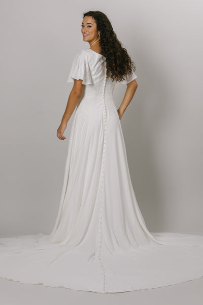 -A backshot of a simple and flowy modest wedding dress that includes a faded lace bodice, flutter sleeves. a soft v-neckline, a synched waist, and a flowy crepe skirt. The view showcases the buttons that go all the way down the backside of the dress.