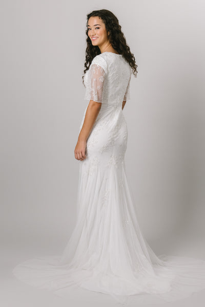 A backshot of a modest wedding dress that features a beaded applique that fades into the skirt and flutter sleeves. It also features a soft scoop neckline and a slim, fitted silhouette.