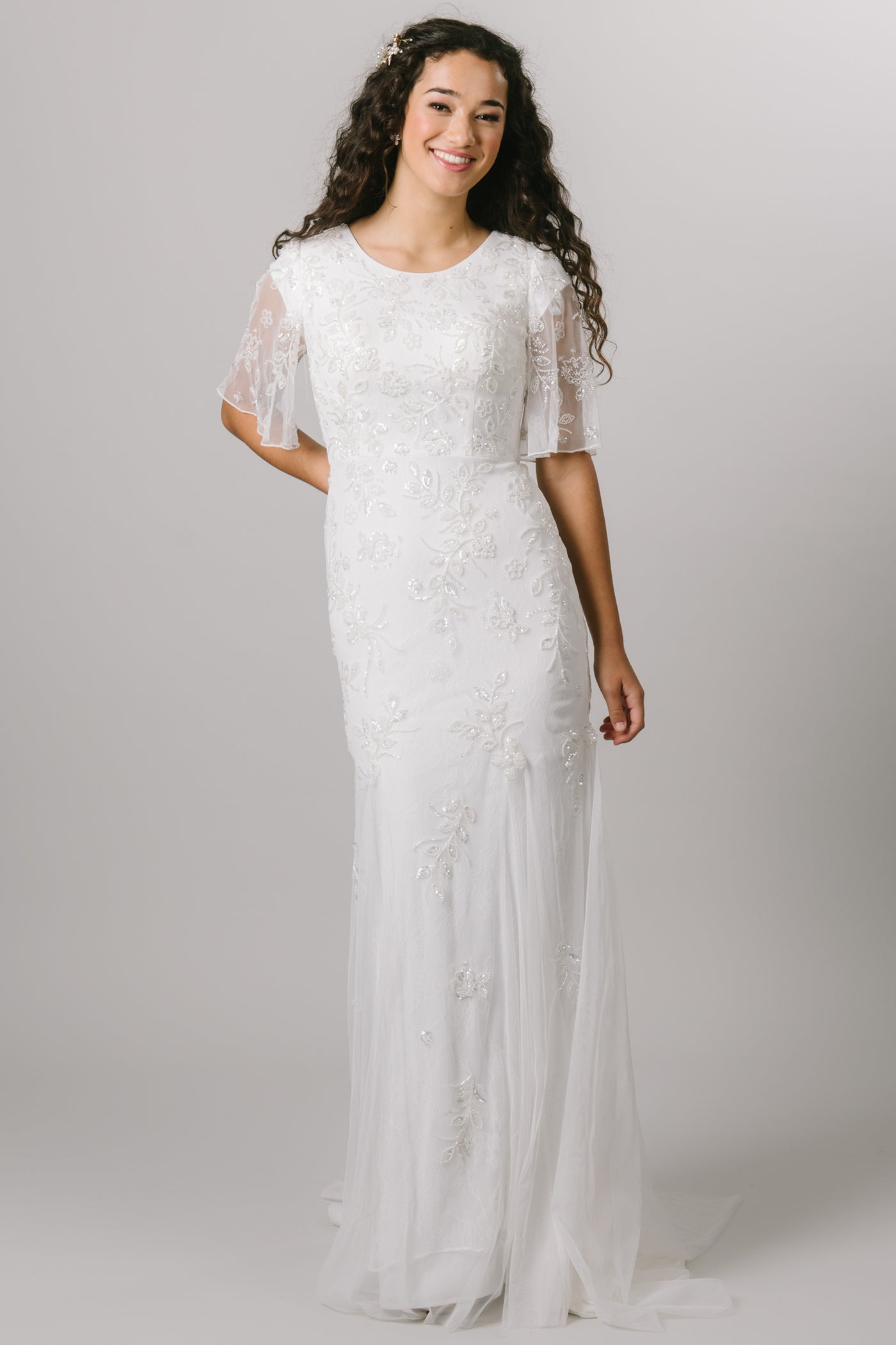This modest wedding dress features a beaded applique that fades into the skirt and flutter sleeves. It also features a soft scoop neckline and a slim, fitted silhouette.