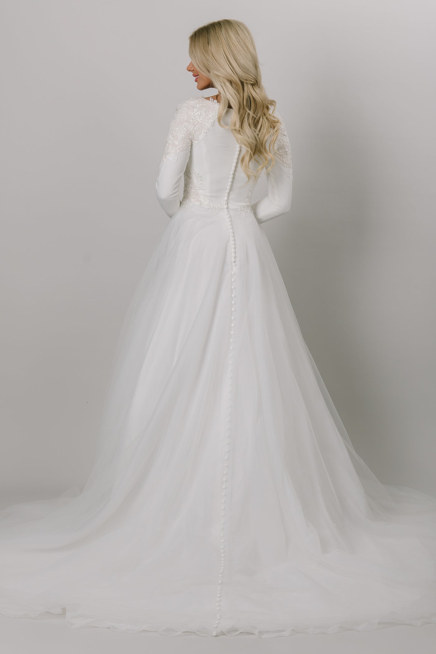Back view of this Moments Made Bridal wedding dress with long sleeves. This modest wedding dress has a high scoop neckline and a-lin fit. This is a perfect wedding dress for those cooler months. It has a zipper covered by buttons down the back.