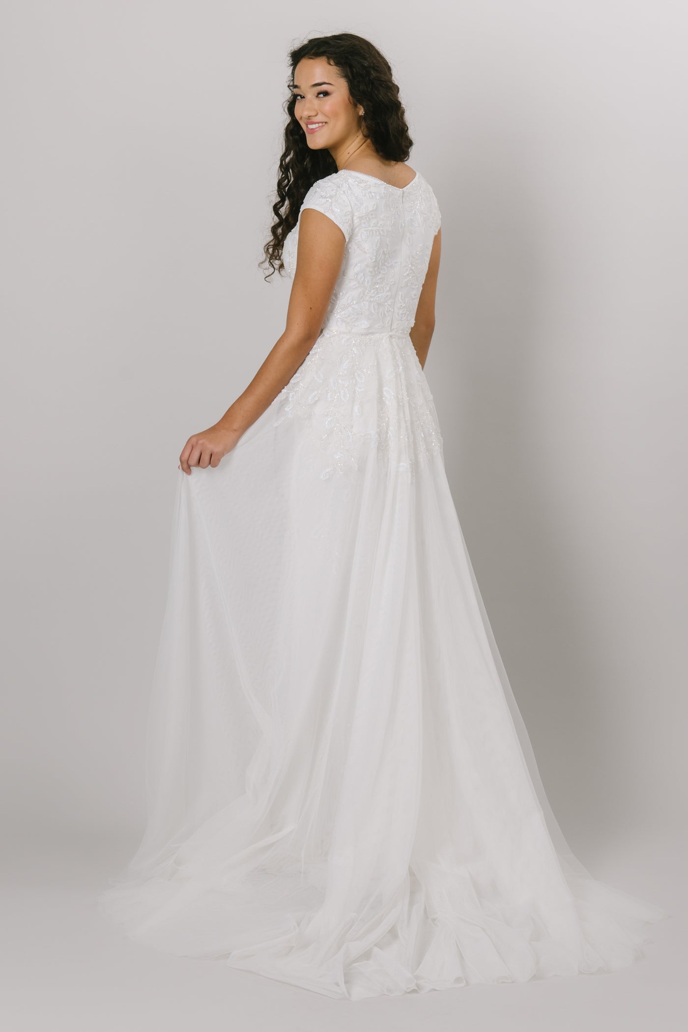 A backshot of the Kalani, which is a fitted, modest wedding dress features a beaded lace applique, cap sleeves, a flowy skirt, and a soft scoop neckline.