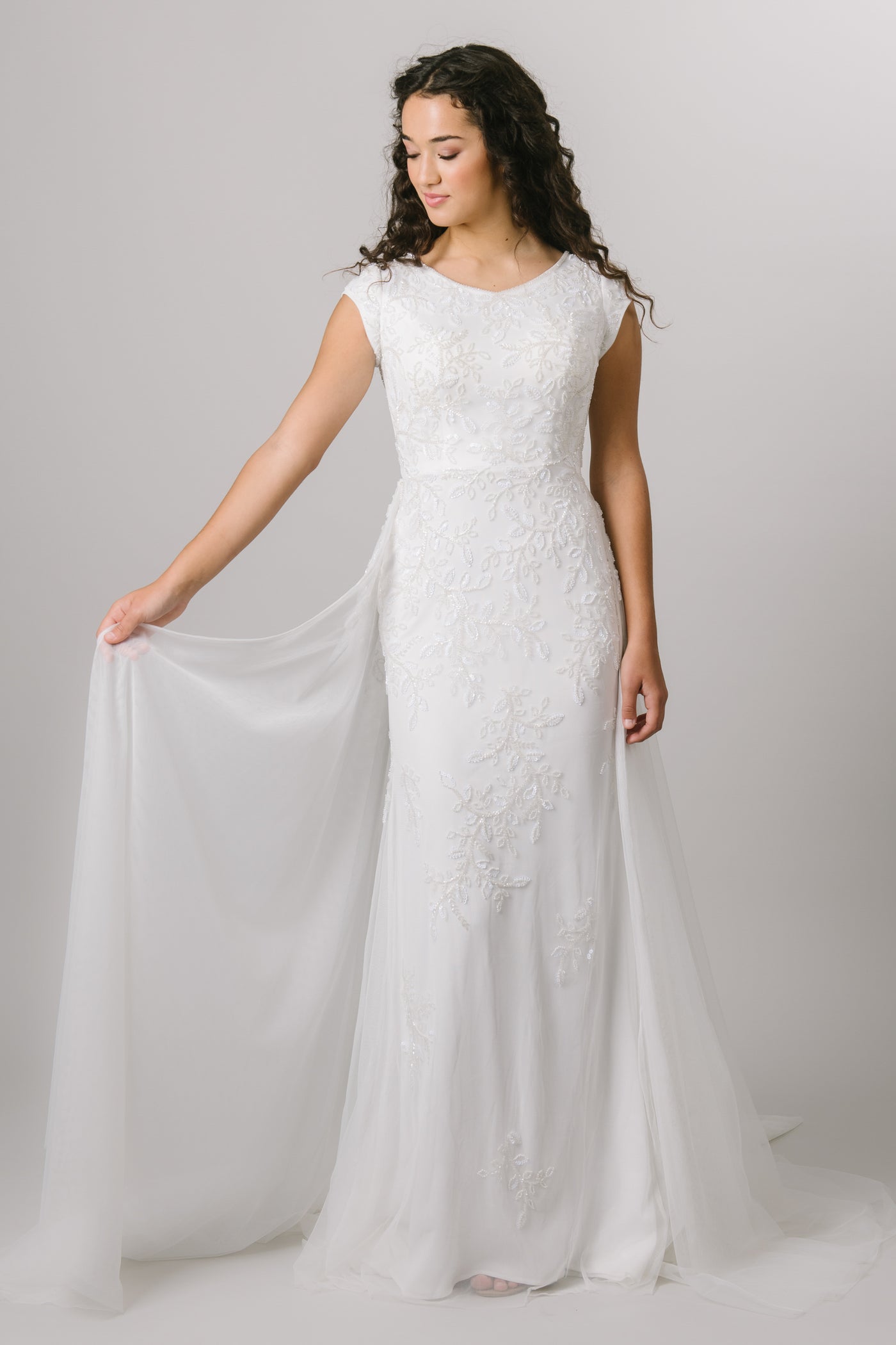The Kalani is a fitted, modest wedding dress features a beaded lace applique, cap sleeves, a flowy skirt, and a soft scoop neckline.