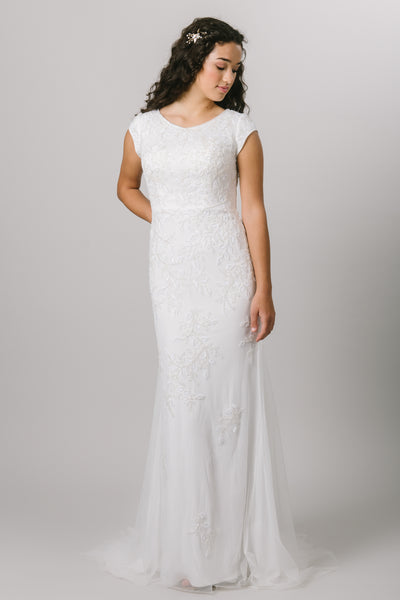 The Kalani is a fitted, modest wedding dress features a beaded lace applique, cap sleeves, and a soft scoop neckline.