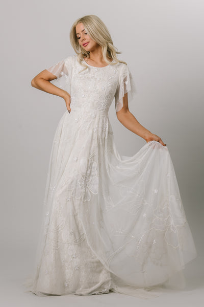 Moments Made Bridal dress with flutter sleeves and a line fit. It is covered in beautiful lace and beading. This modest wedding dress has the pattern has a flower like design.