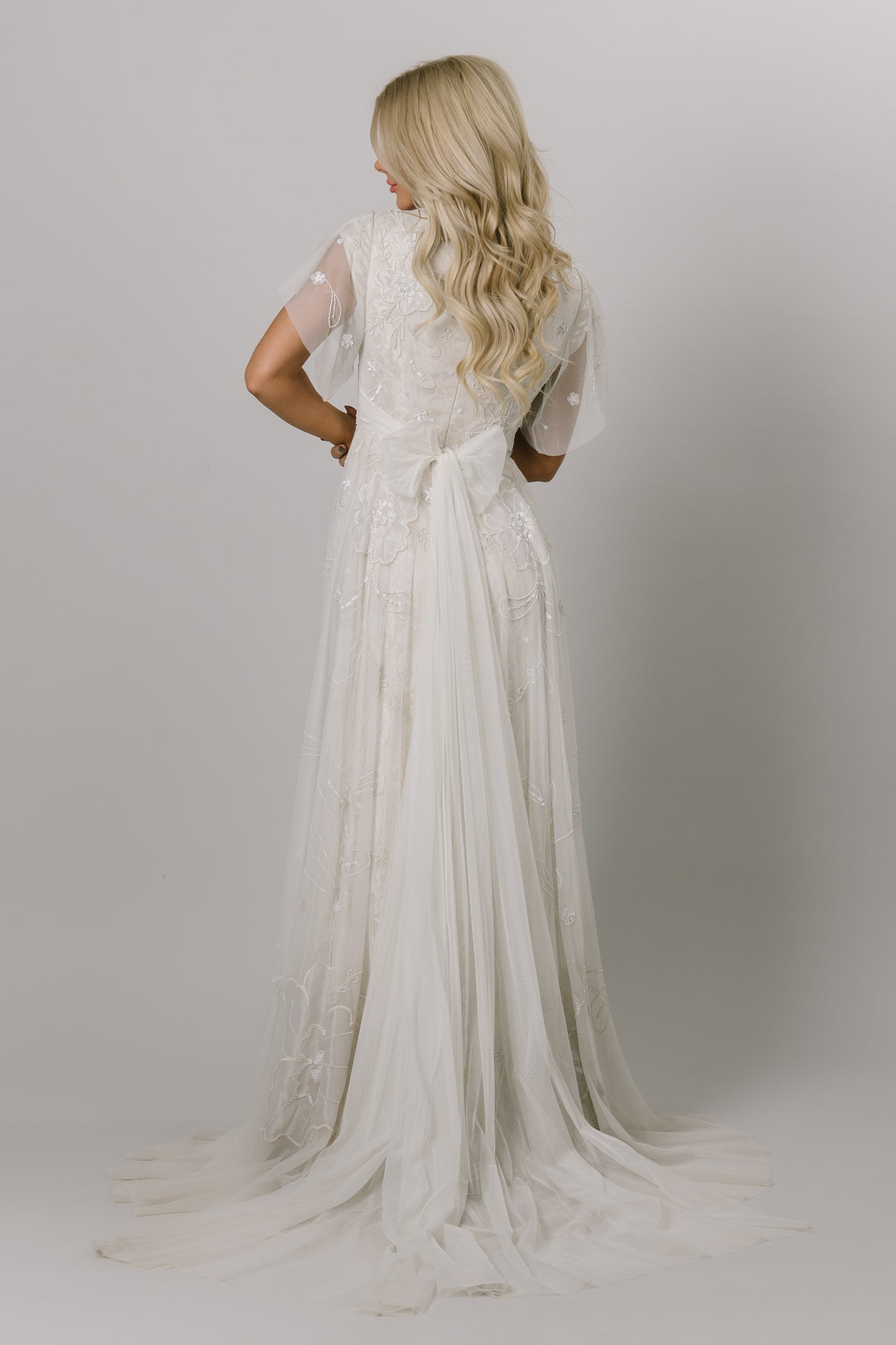 Back view of Moments Made Bridal dress with flutter sleeves and a line fit. It is covered in beautiful lace and beading. This modest wedding dress has the pattern has a flower like design.
