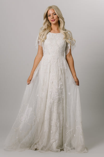 Moments Made Bridal dress with flutter sleeves and a line fit. It is covered in beautiful lace and beading. This modest wedding dress has the pattern has a flower like design.