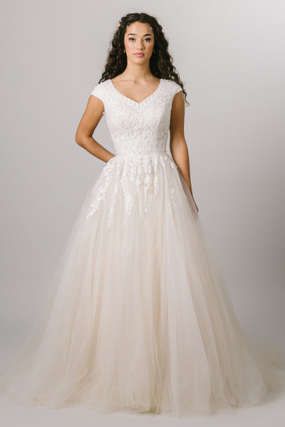 This modest wedding dress features a faded lace bodice, a v-neckline, cap sleeves, a synched waist, and a full tool skirt.