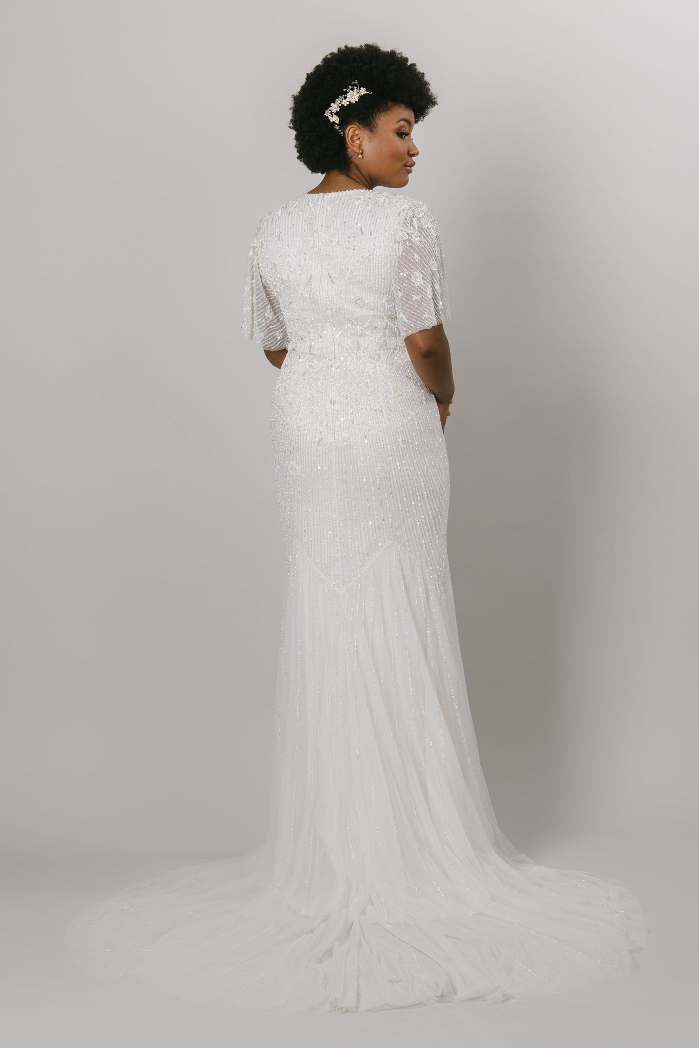 This fully jeweled modest wedding gown features a delicate flutter sleeve, and soft mermaid silhouette. Available in ivory.