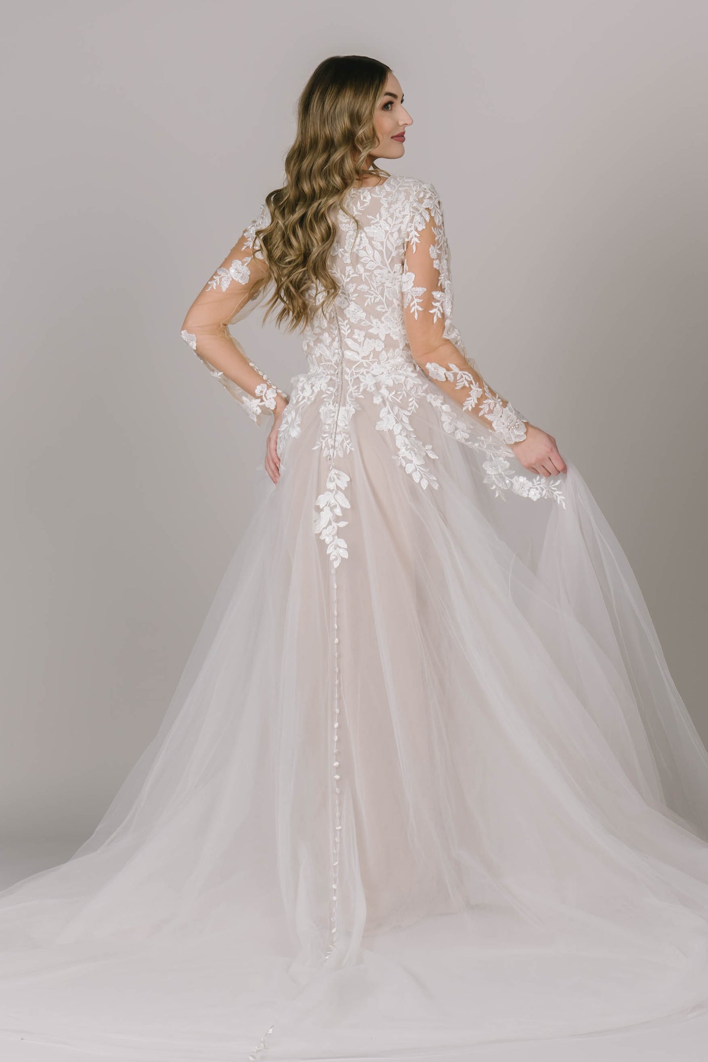 A back shot of this modest wedding dress with buttons downs the back of the dress, sheer long sleeves with applique, and a-line silhouette.