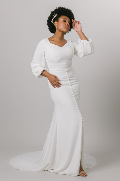 A fitted modest wedding dress which features include a simple crepe fabric that is synched at the waist, buttons going down the front, a knee high slit, and bishop sleeves.