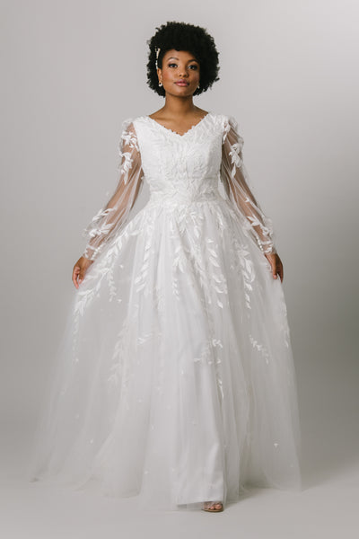 A modest ballgown wedding dress that features a lace bodice that fades out through the tool skirt, v-neckline, and long, sheer, bishop sleeves.
