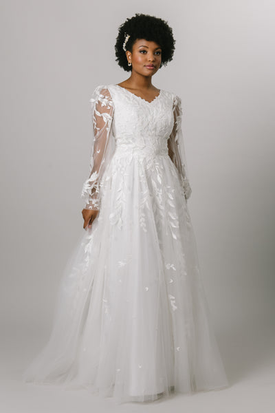 A modest ballgown wedding dress that features a lace bodice that fades out through the tool skirt, v-neckline, and long, sheer, bishop sleeves.