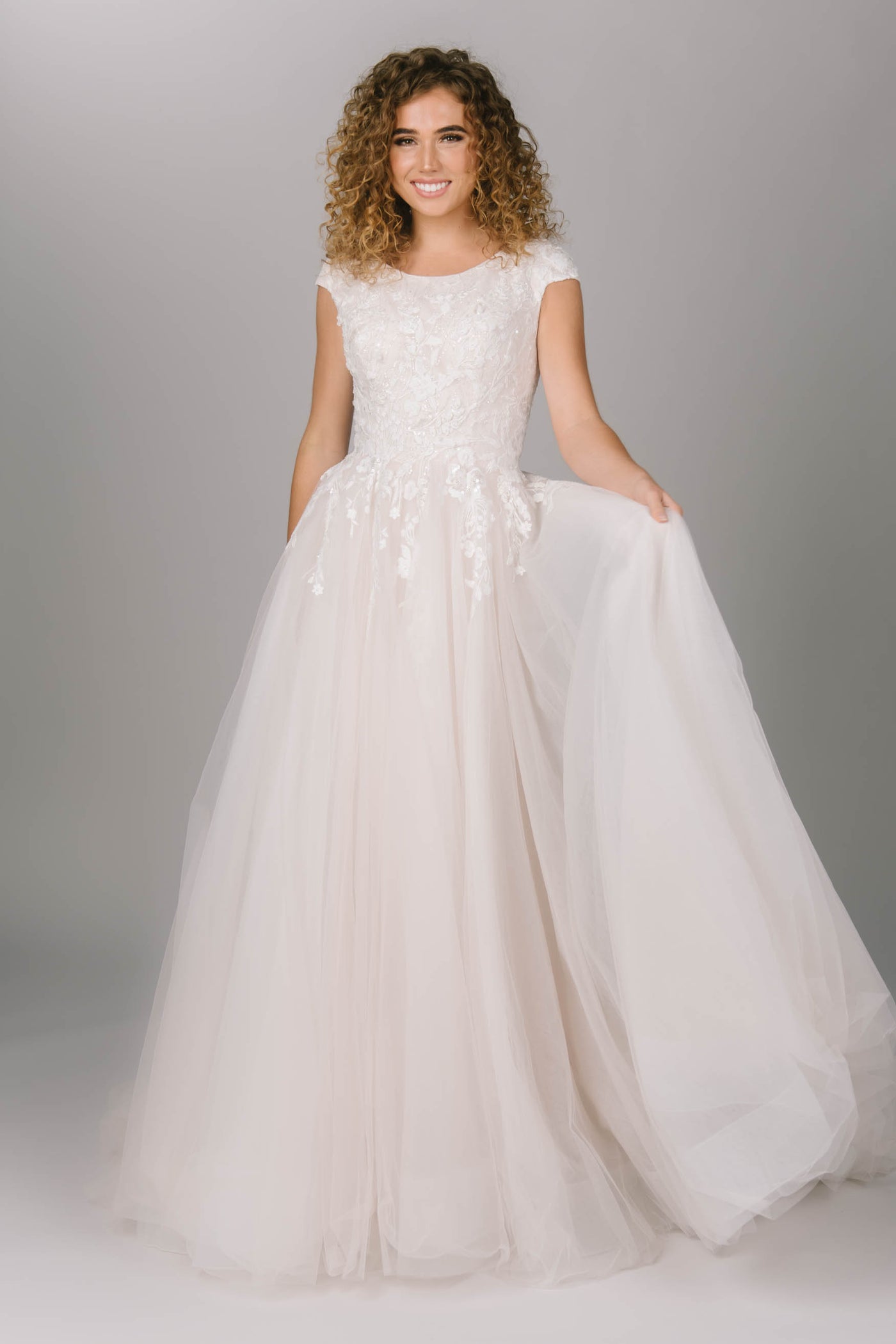 Front view of modest ballgown wedding dress. This dress has cap sleeves and a tulle bottom. It has delicate lace trickling down the waistline of the dress. It has a zipper with buttons all down the train. Magical modest ballgown wedding dress. 