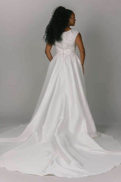 Modest wedding dress with v-neckline and cap sleeves. It has a lace top that trickles down the waistline of the dress. It has a longer train. Buttons run all the way down the dress. It is a stunning and simple modest wedding dress.