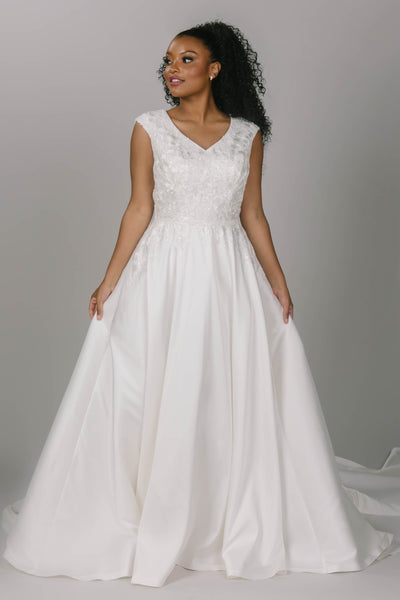 Modest wedding dress with v-neckline and cap sleeves. It has a lace top that trickles down the waistline of the dress. It has a longer train. Buttons run all the way down the dress. It is a stunning and simple modest wedding dress. 