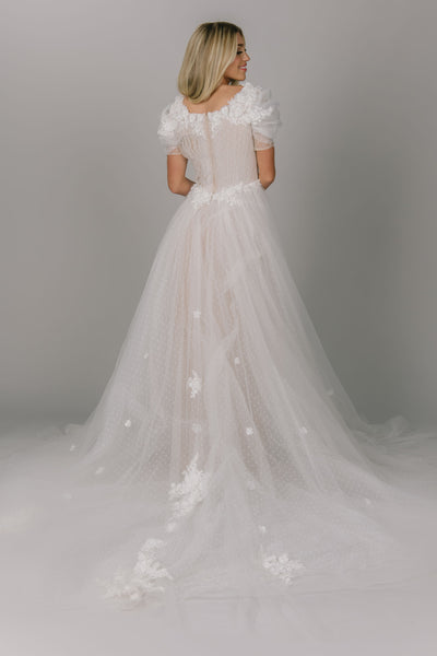 Dreamy modest wedding dress with puffed sleeves. It has a 3D flower lace that lines the scoop neckline. It has a slit and a lower train. It has multiple layers of tulle, of which one has polka dots.