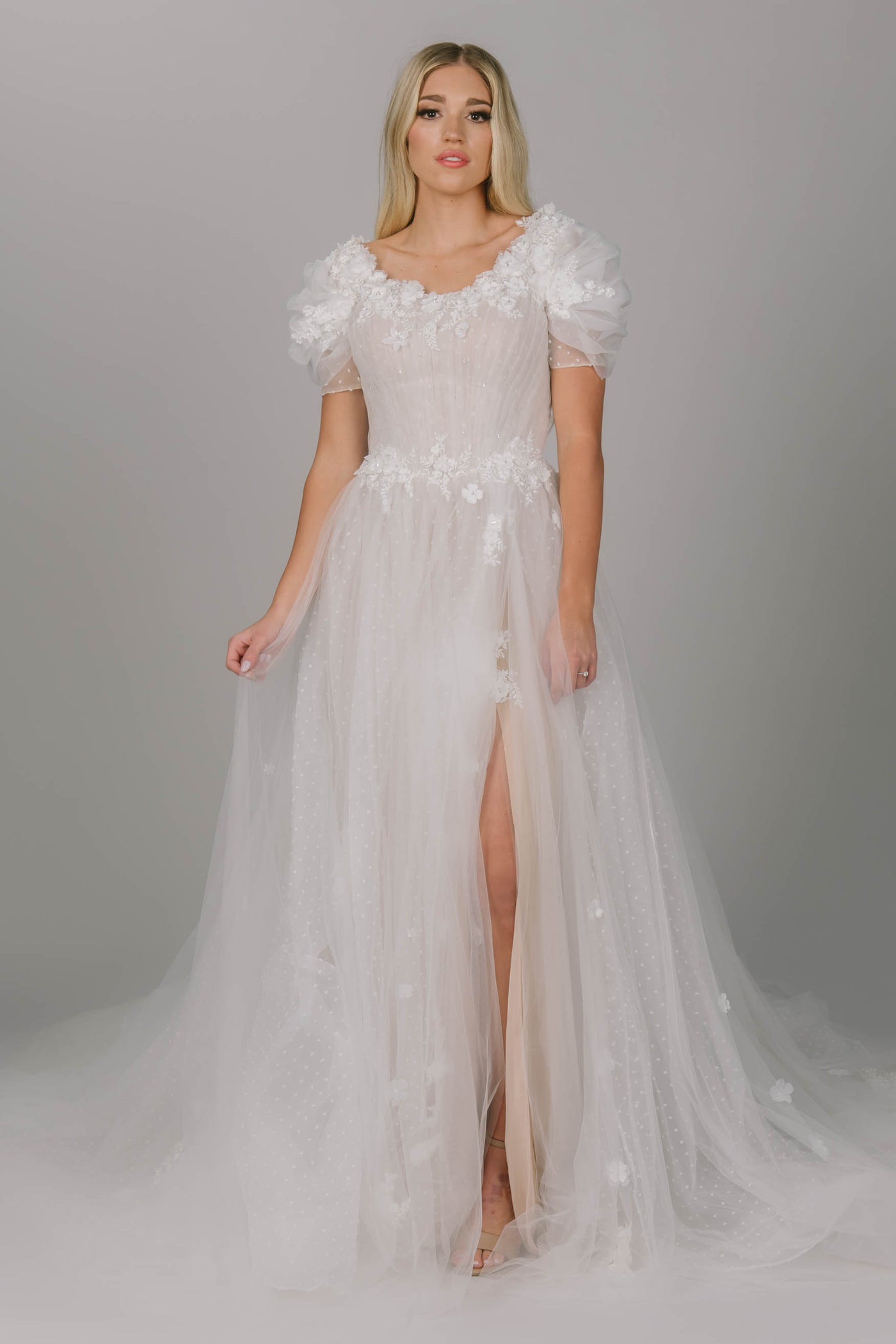 Dreamy modest wedding dress with puffed sleeves. It has a 3D flower lace that lines the scoop neckline. It has a slit and a lower train. It has multiple layers of tulle, of which one has polka dots. 