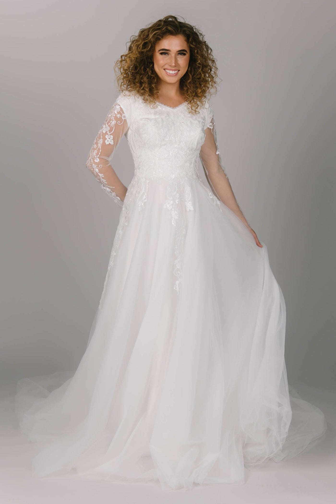 Front view of modest wedding dress with vine-like lace. It is a-line with a v-neckline. It has a beautiful tulle skirt and long sleeves. It is a dreamy modest wedding dress. 