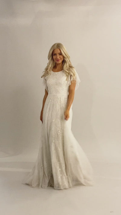 Video of Moments Made Bridal dress with flutter sleeves and a line fit. It is covered in beautiful lace and beading. This modest wedding dress has the pattern has a flower like design.