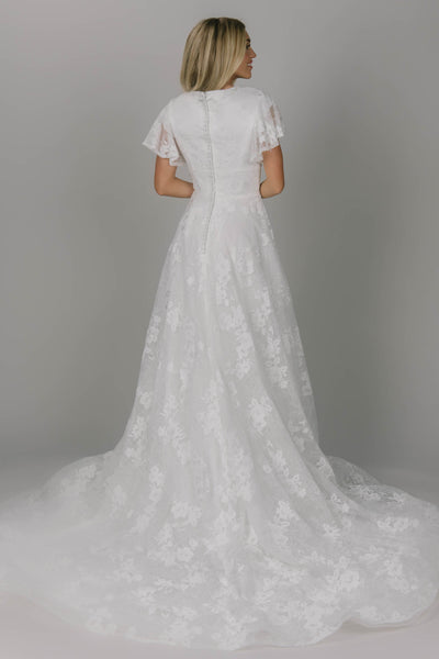 Modest wedding dress with a-line fit. It has flutter sleeves and a high scoop neckline. It has covered buttons on the back. It is ivory with a flower sheet lace.