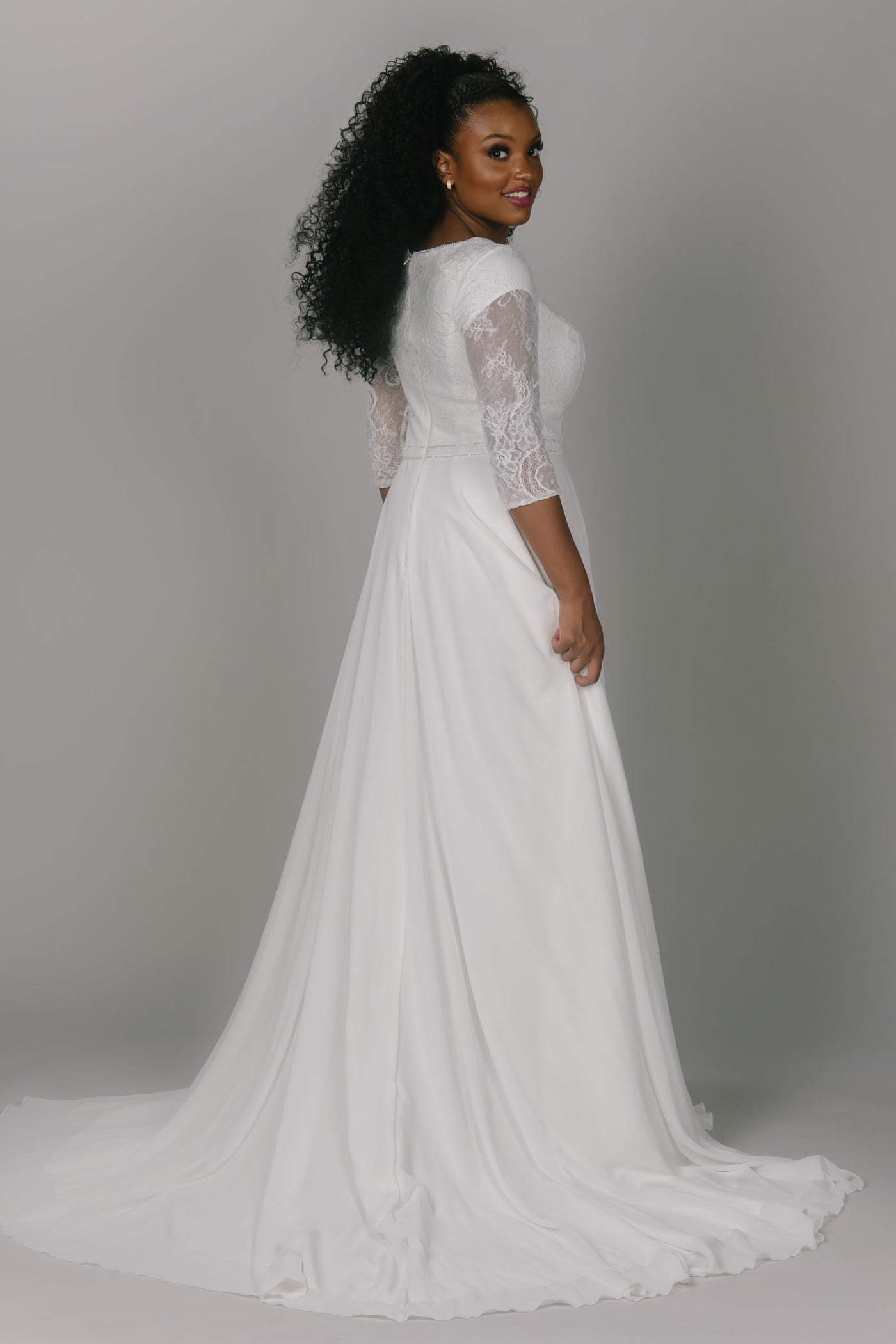 Modest wedding dress featuring a lace top and chiffon bottom. It has a high scoop neckline and three quarter length sleeves. It has a sparkly belt and a flowy skirt. This modest wedding dress is an icon for this particular style.