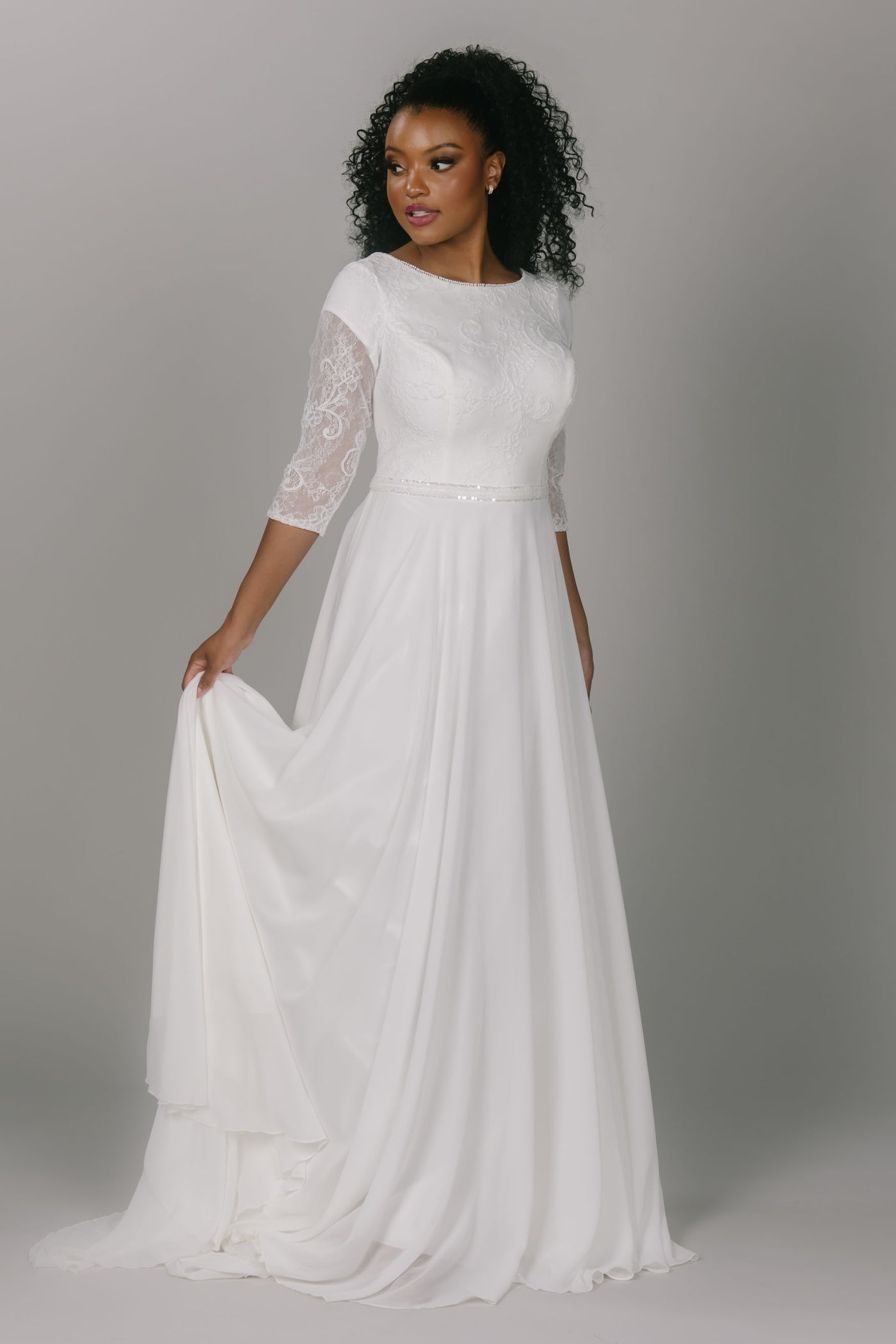 Modest wedding dress featuring a lace top and chiffon bottom. It has a high scoop neckline and three quarter length sleeves. It has a sparkly belt and a flowy skirt. This modest wedding dress is an icon for this particular style. 