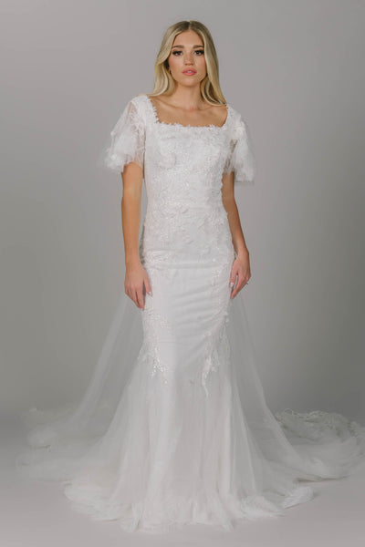 Sparkly modest wedding dress featuring a square neckline and flutter sleeves. It is a 3D lace and beaded fitted gown. It has a longer train. This view shows the overskirt that can be sold separately.