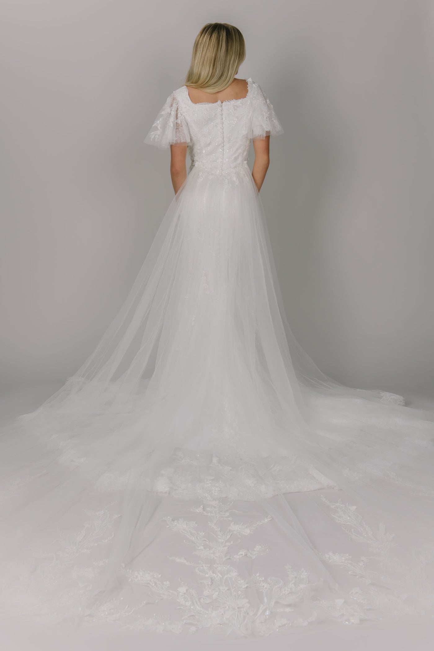 Sparkly modest wedding dress featuring a square neckline and flutter sleeves. It is a 3D lace and beaded fitted gown. It has a longer train. This view shows the overskirt that can be sold separately.