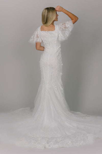 Sparkly modest wedding dress featuring a square neckline and flutter sleeves. It is a 3D lace and beaded fitted gown. It has a longer train with buttons down the back. 